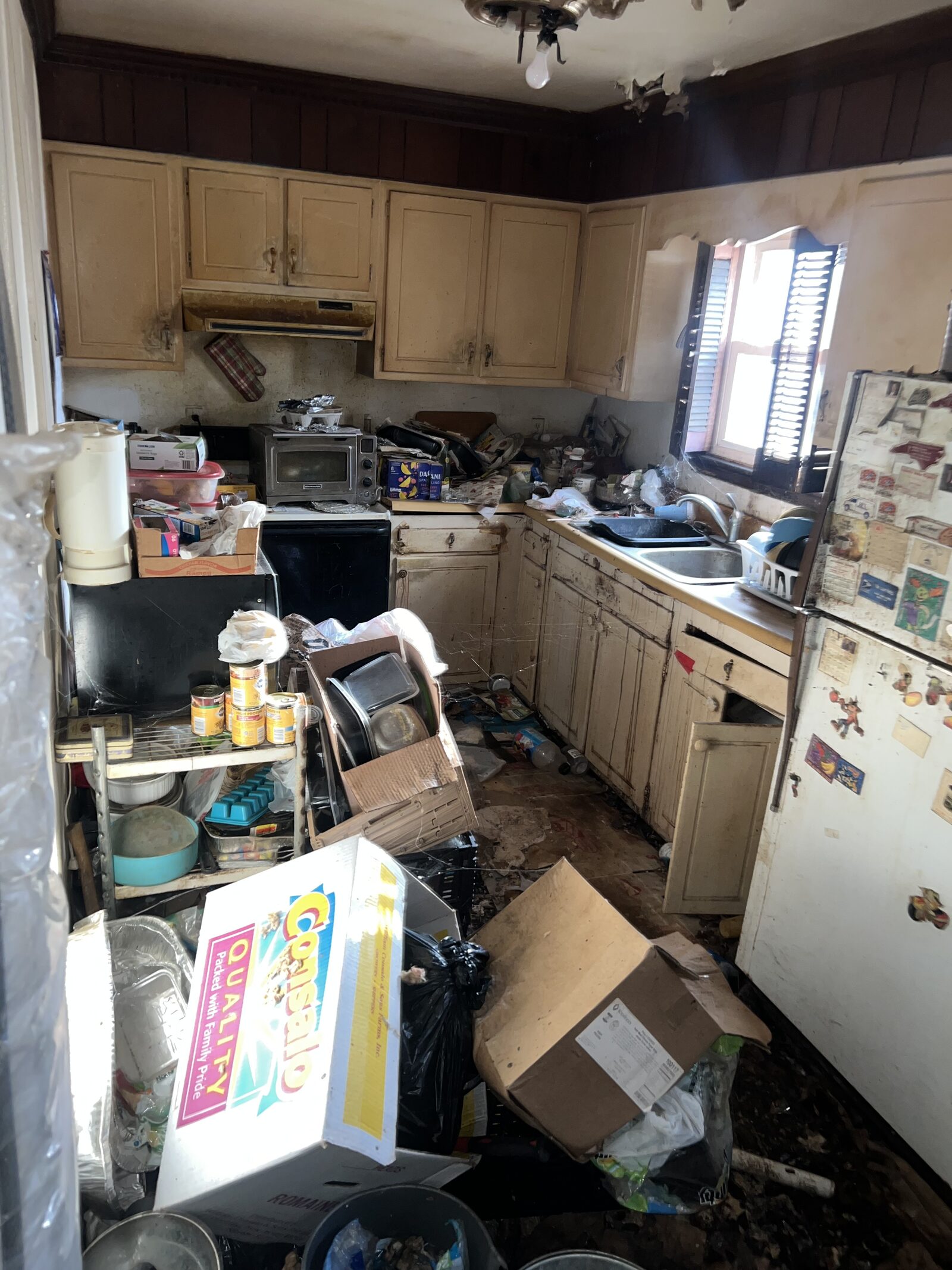 Severely cluttered kitchen with damaged cabinets, peeling ceiling and debris on the floor