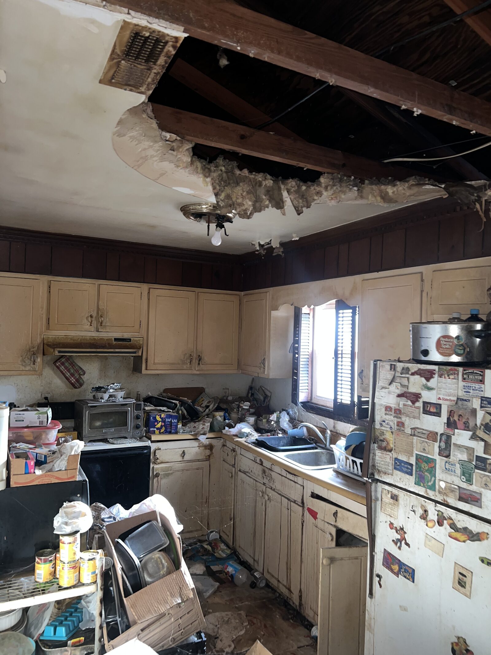 Kitchen with a collapsed ceiling exposing rafters above worn cabinets and clutter